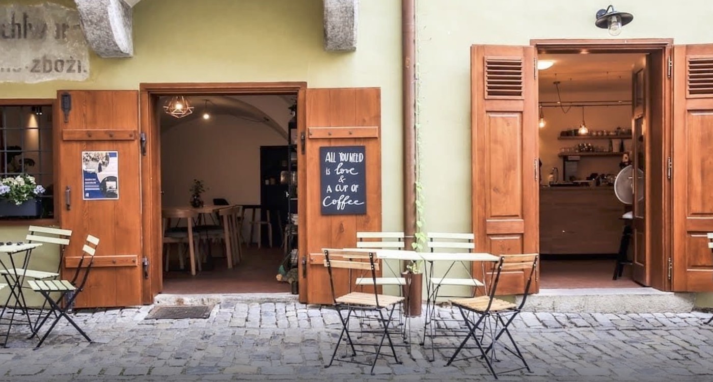 The open wooden shutter doors of Masná 130 with outdoor cafe tables on the cobblestones and a chalkboard sign reading All you need is love and a cup of coffee